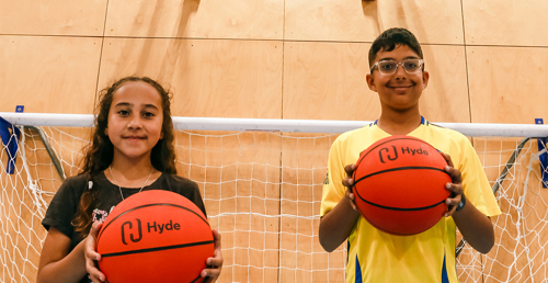 Two of Young Harrow Foundation’s members, a girl and a boy, smiling and each holding a basketball with the Hyde name and logo on.